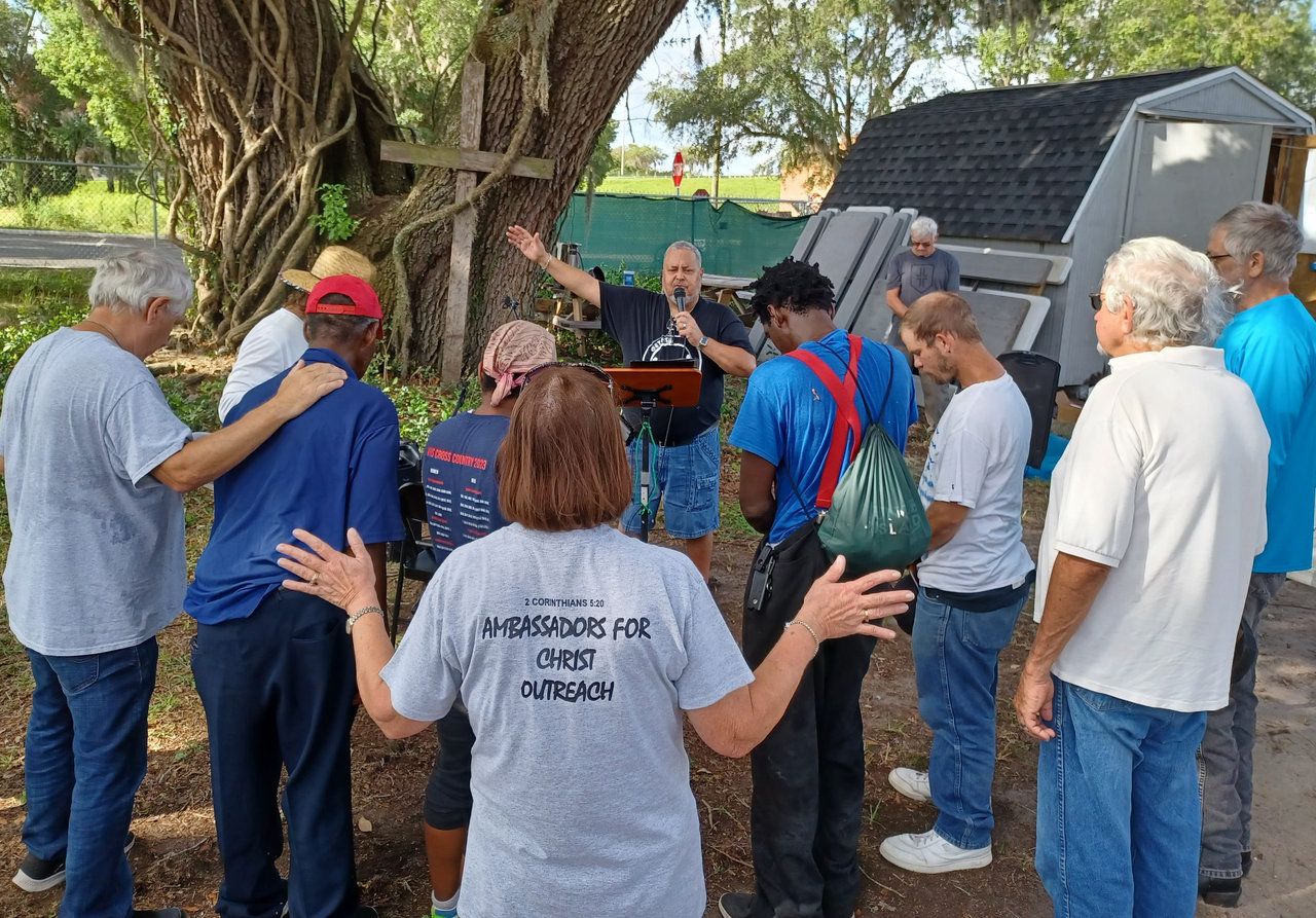 An AFCO 5:20 leader preaches at an outdoor altar call beneath a wooden cross, one arm raised, as neighbors bow their heads and lay hands on one another’s shoulders.