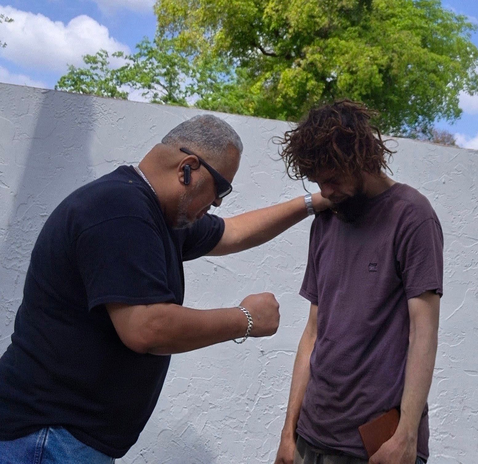 An AFCO 5:20 volunteer prays with a young man outside, one hand resting on his shoulder as both bow their heads.